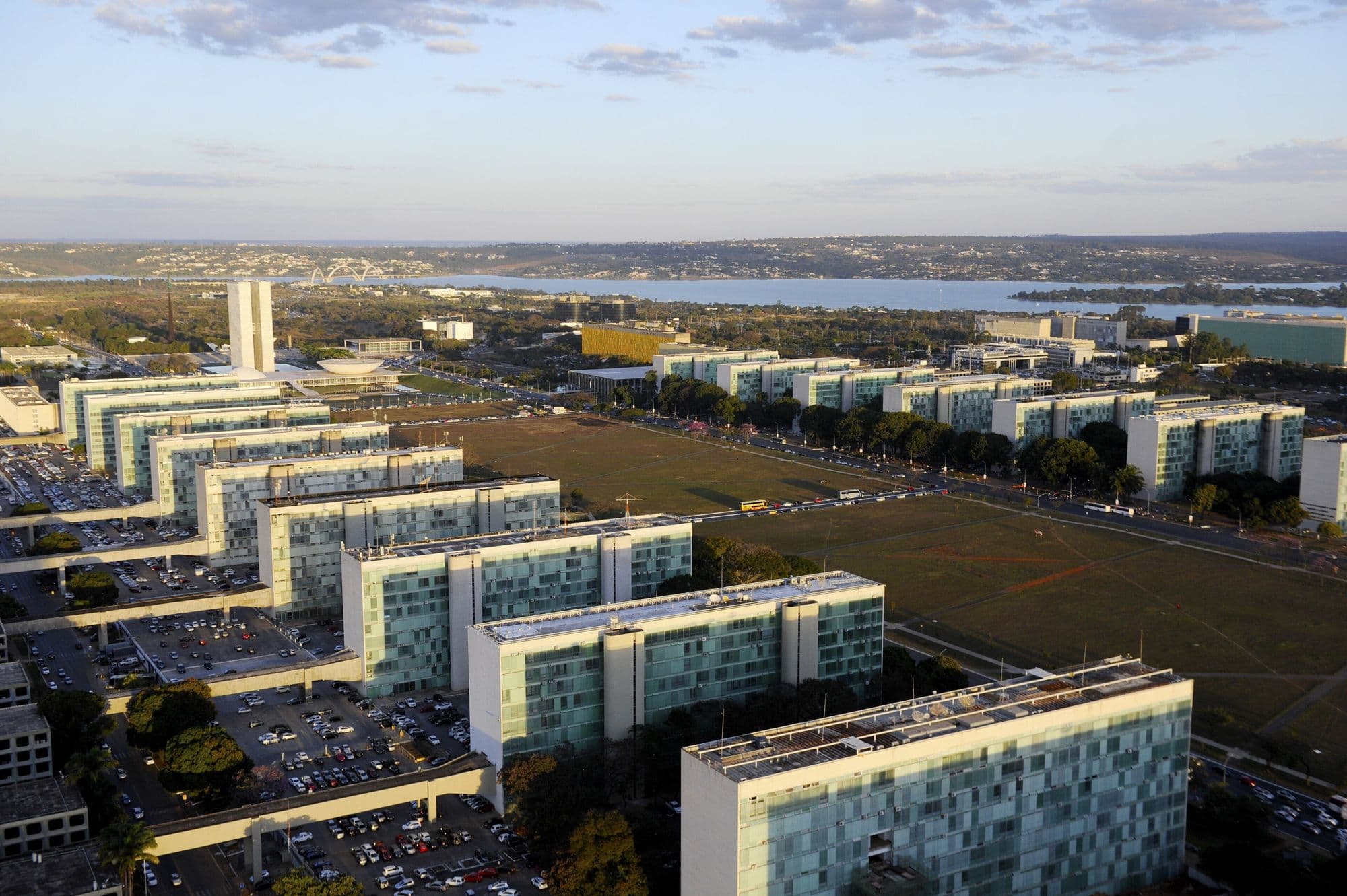 Blocos dos ministérios na Esplanada, vistos do alto, com o Congresso e o Lago Paranoá ao fundo.